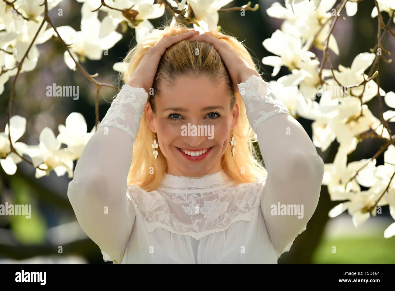 Blonde woman driving her hands through her hair, portrait in front of a blooming magnolia tree, Baden-Wurttemberg, Germany Stock Photo