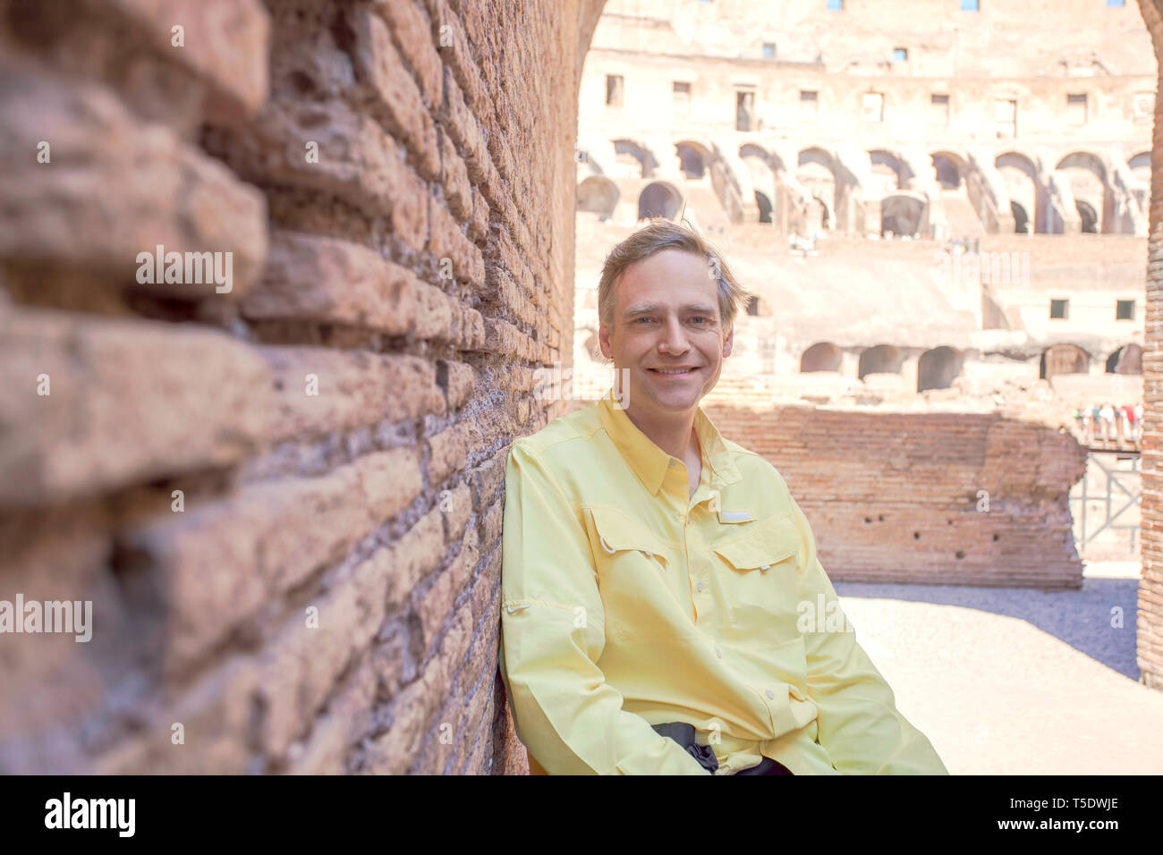 Smiling Caucasian man in yellow shirt, sitting and leaning against ancient brick wall  inside the Colloseum in Rome, Italy Stock Photo