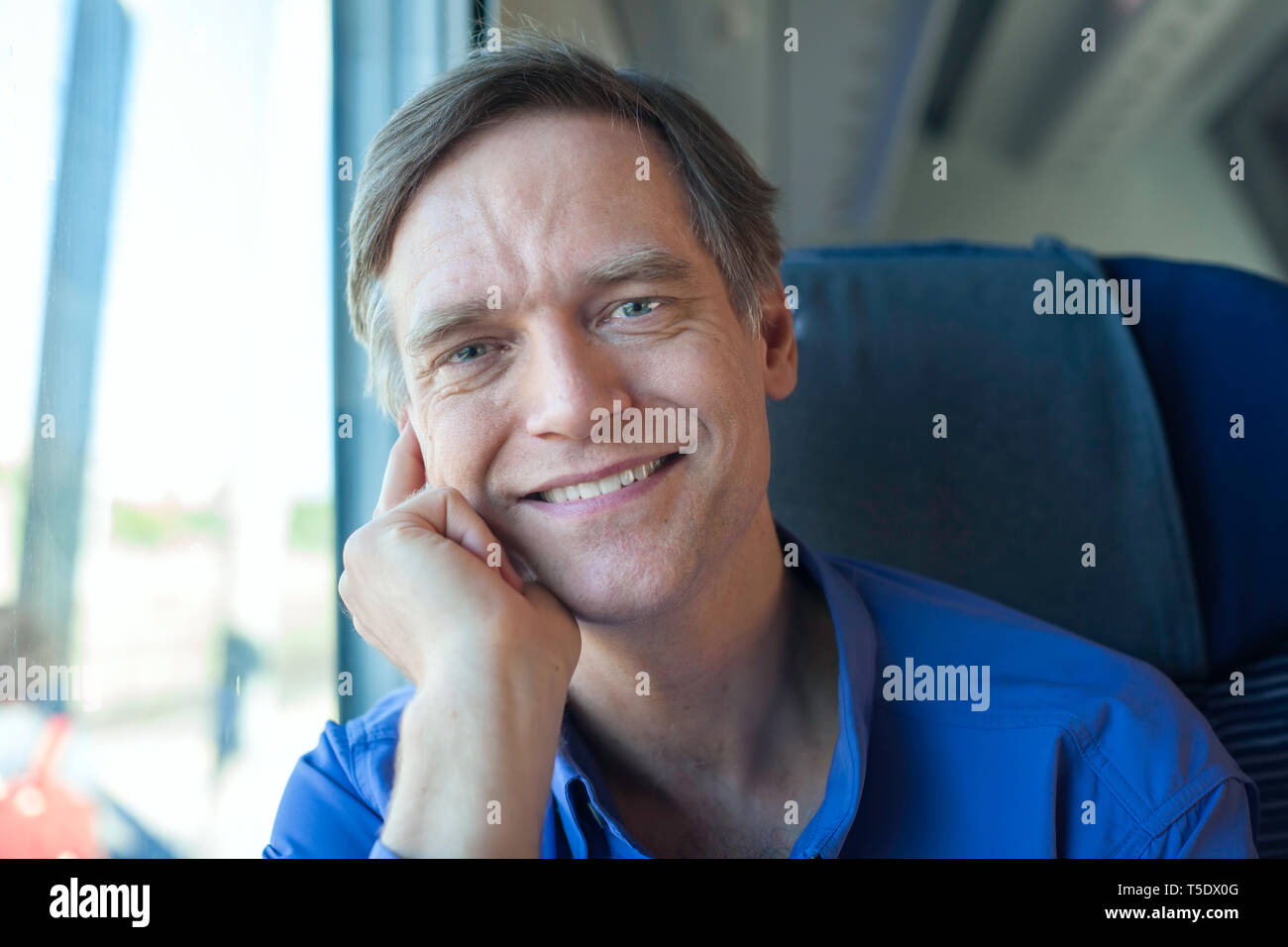 handsome Caucasian man in blue shirt sitting on commuter train in daytime, smiling at camera Stock Photo
