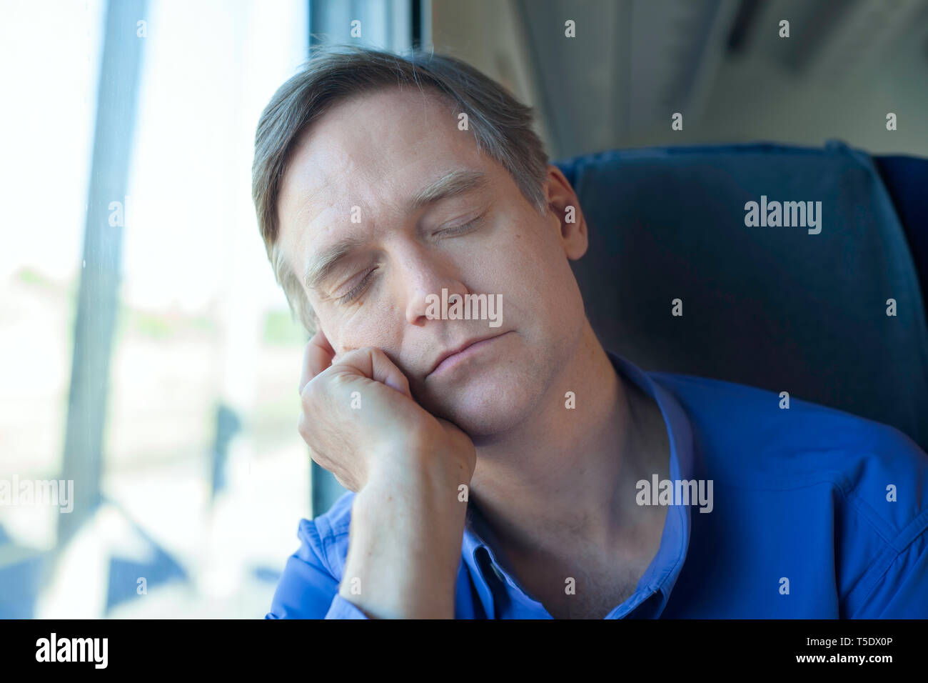 Caucasian man wearing blue shirt in early fifties sleeping and resting head on hand near train window Stock Photo