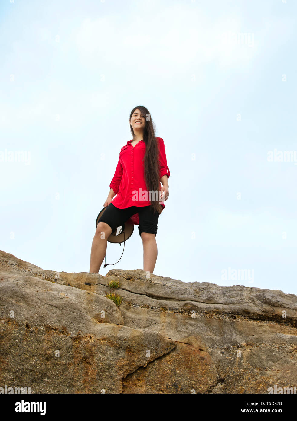 Biracial Caucasian  Asian teen girl with long hair in red shirt standing on top of high rocky summit smiling down on camera Stock Photo