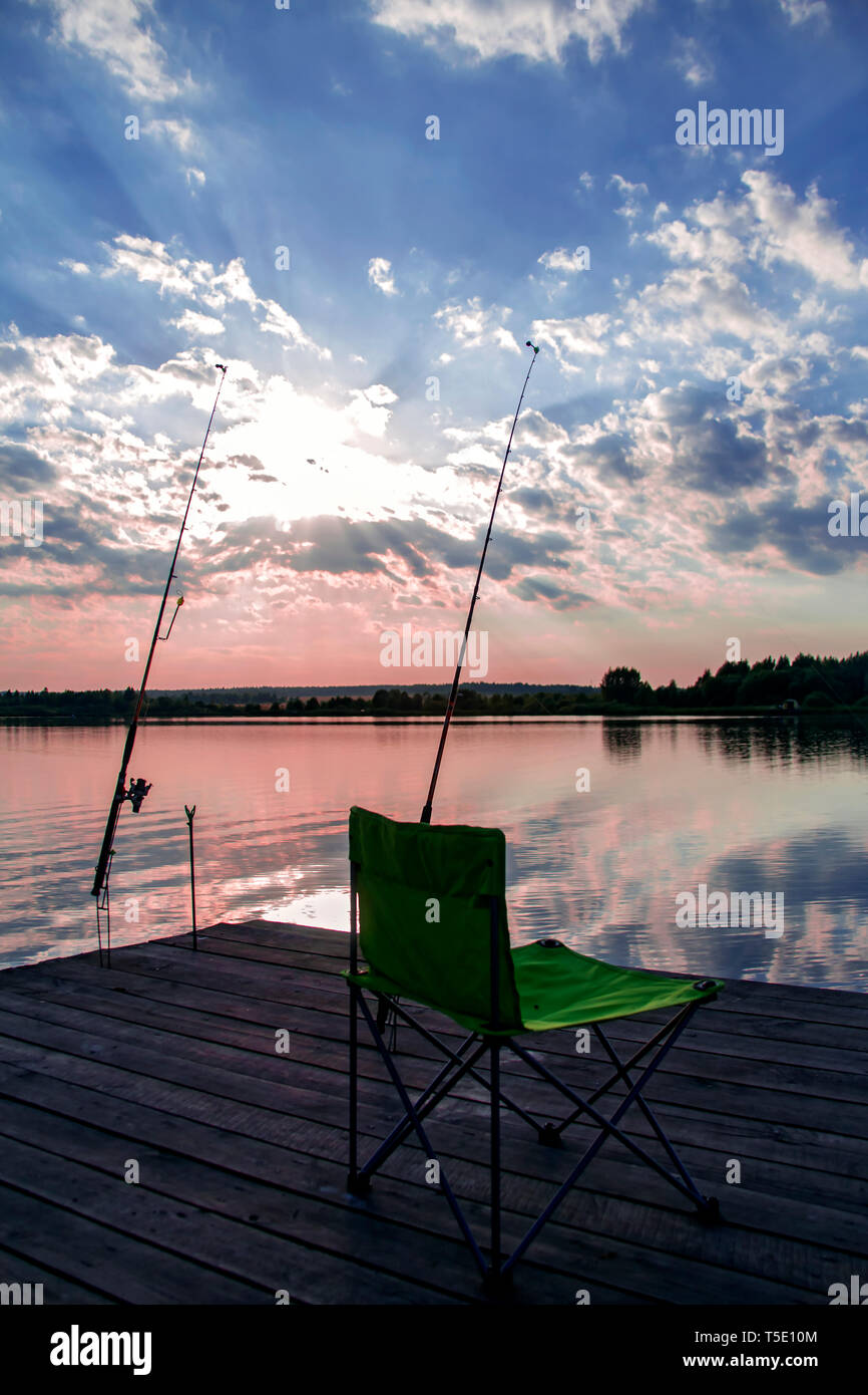 Folding fishing chair and two fishing rods on a wooden pontoon on the lake at sunset Stock Photo