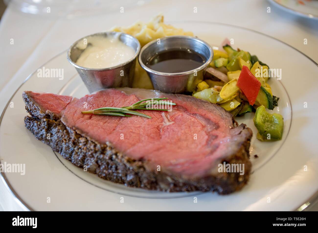 A cut of Prime Rib beef on a white plate with sides, horse radish and Au Jus shot shallow depth of field with focus on the rosemary sprig. Stock Photo