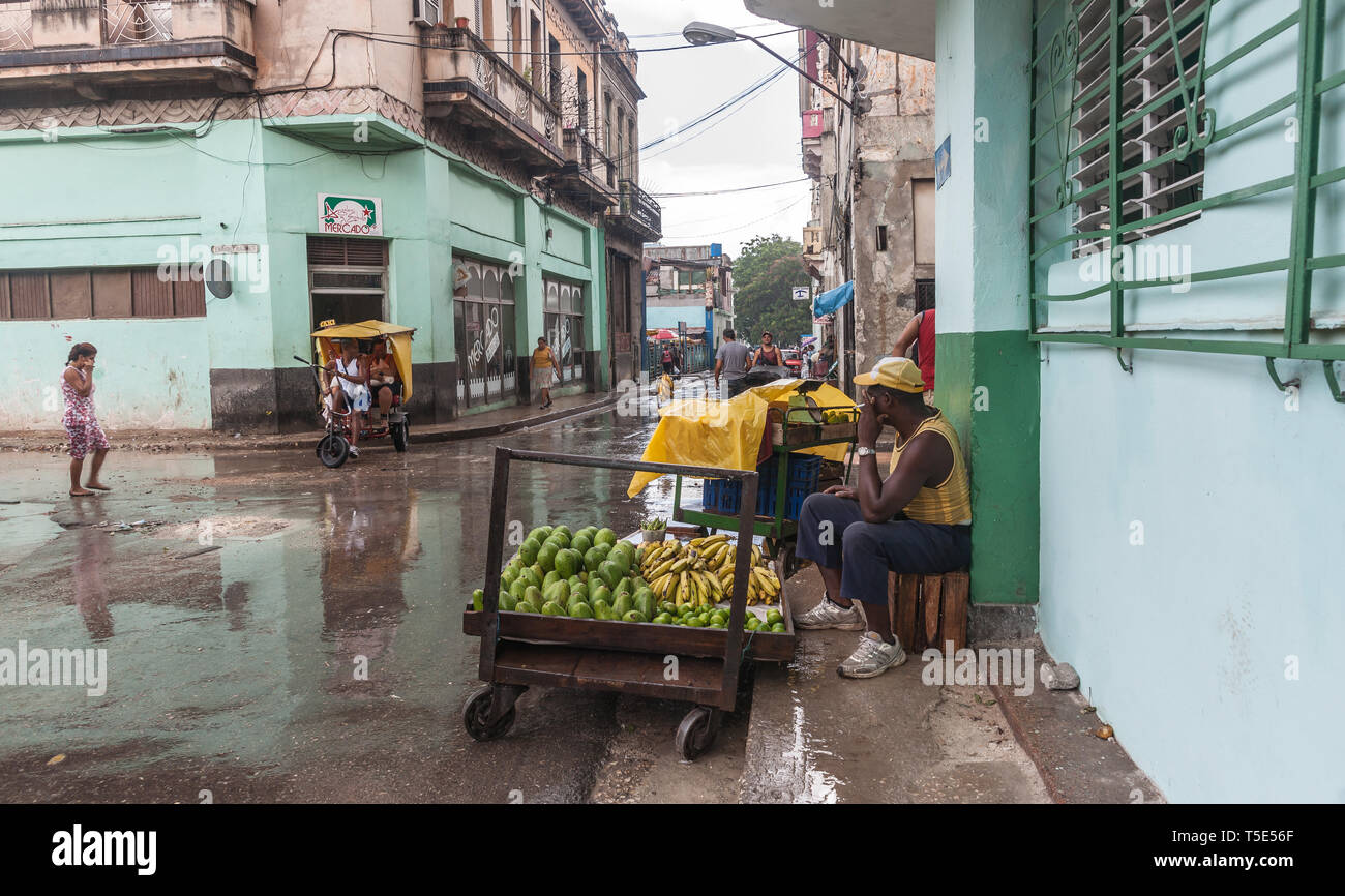 HAVANA CUBA - JULY 8 2012; Heavy rain in street of Havana with man and fruit cart sits sheltering from heavy rain hoping to sell while others walk on  Stock Photo
