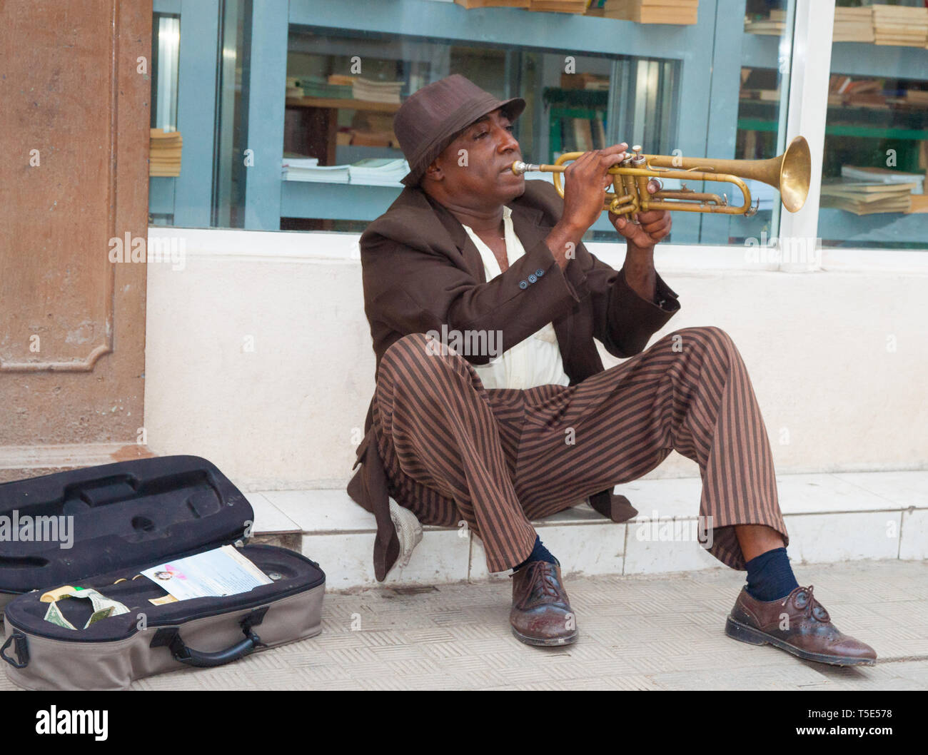 HAVANA CUBA - JULY 8 2012; Street music  in Havana with Afro-Cuban man busking while sitting on footpath in front shop window playing trumpet. Stock Photo