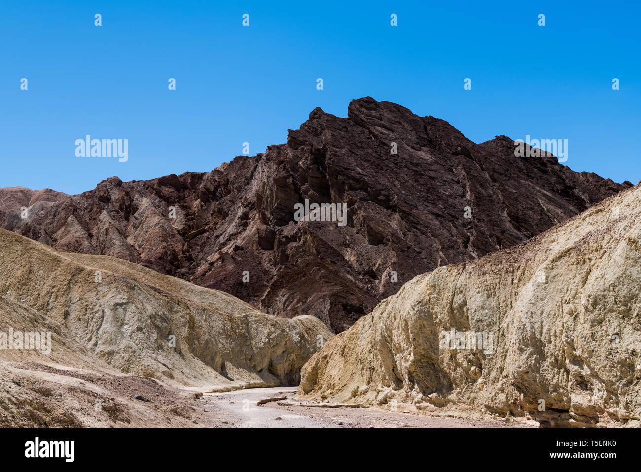 A dry riverbed cuts a pathway through a colorful desert canyon landscape to a rugged mountain peak - Golden Canyon in Death Valley National Park Stock Photo