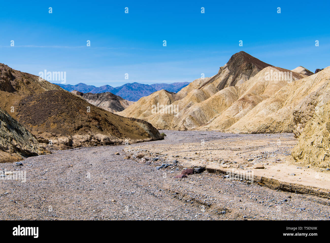 A dry rocky riverbed curves through a barren desert landscape of colorful badlands and peaks in Death Valley National Park Stock Photo
