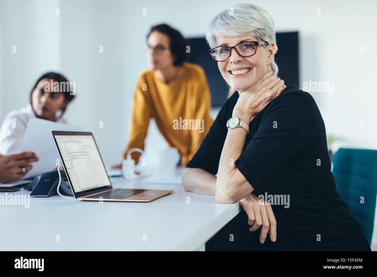Successful mid adult woman sitting at a meeting with colleagues in boardroom. Female with coworkers in conference room looking at camera and smiling. Stock Photo