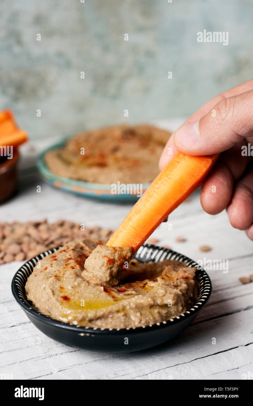 closeup of a man dipping a strip of carrot in a homemade lentil hummus seasoned with paprika served in a green ceramic plate, on a white rustic wooden Stock Photo
