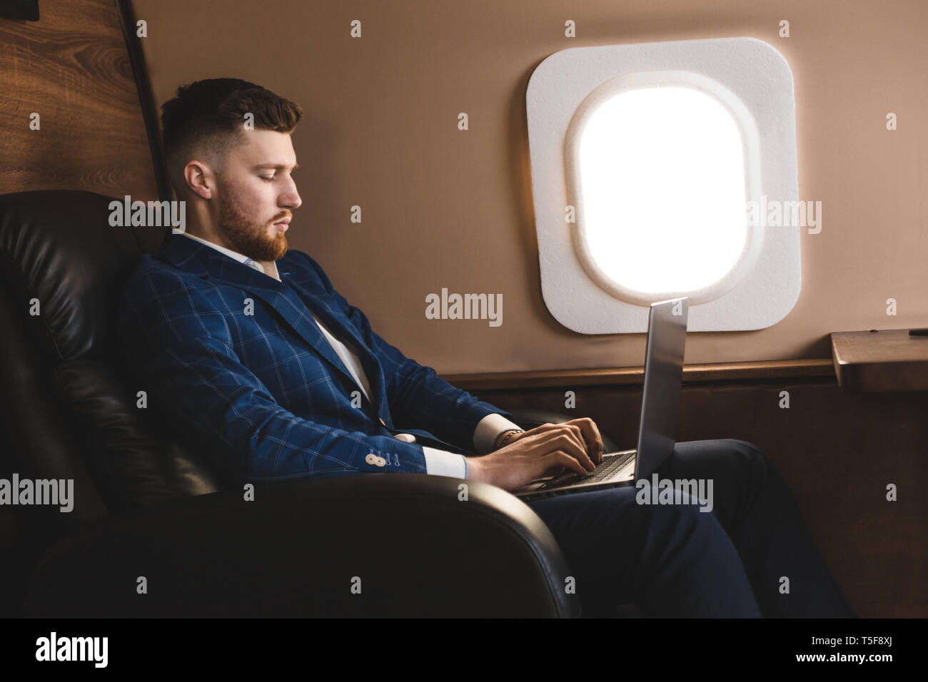 Attractive and successful businessman working on a laptop while sitting in a chair of his private jet. Stock Photo