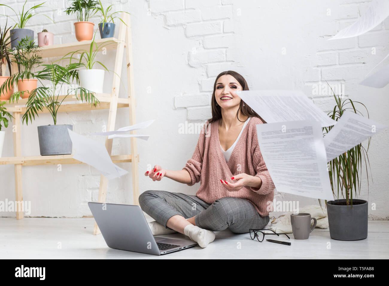 A young happy woman throws up a bunch of pieces of paper and documents sitting near the laptop, the freelance girl is happy and in a good mood Stock Photo