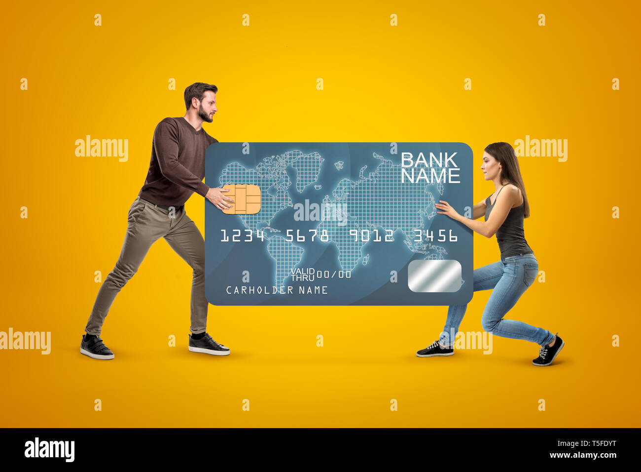 Young man and woman in casual clothes carrying a big plastic bank card on yellow background Stock Photo