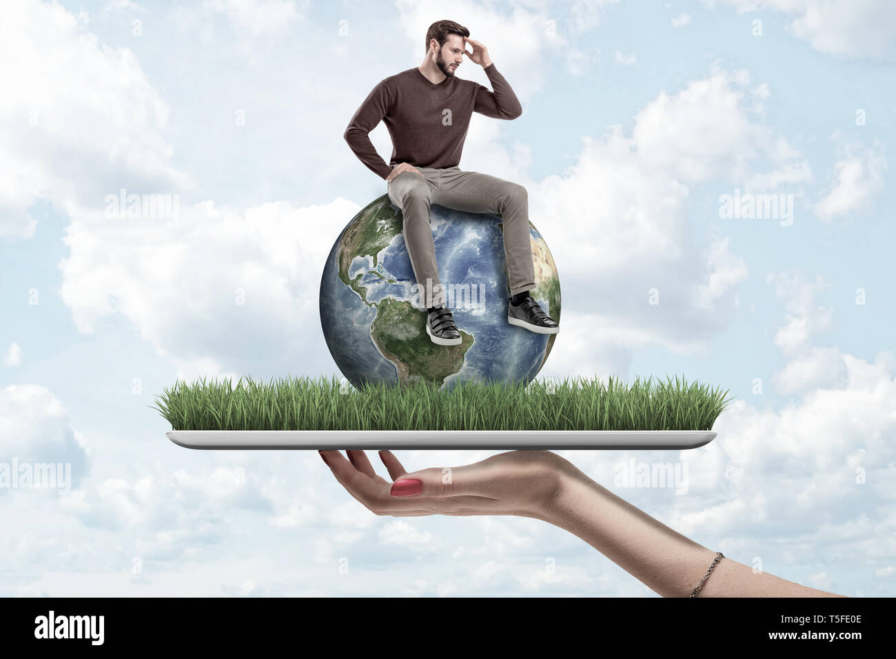 Female hand holding small man in casual clothes sitting on earth globe on green grass model with blue sky background Stock Photo