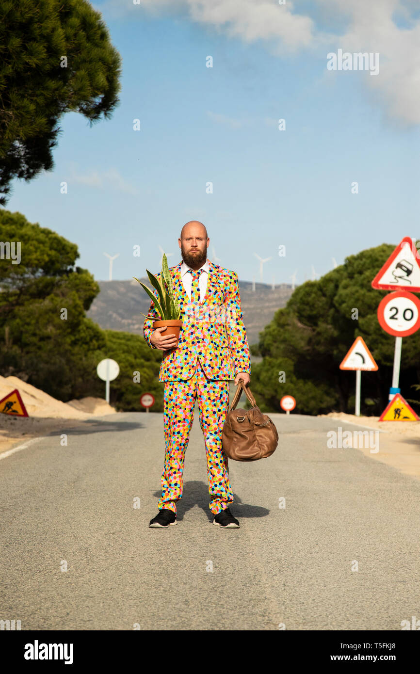 Man wearing suit with colourful polka-dots standing on country road with travelling bag and potted plant Stock Photo