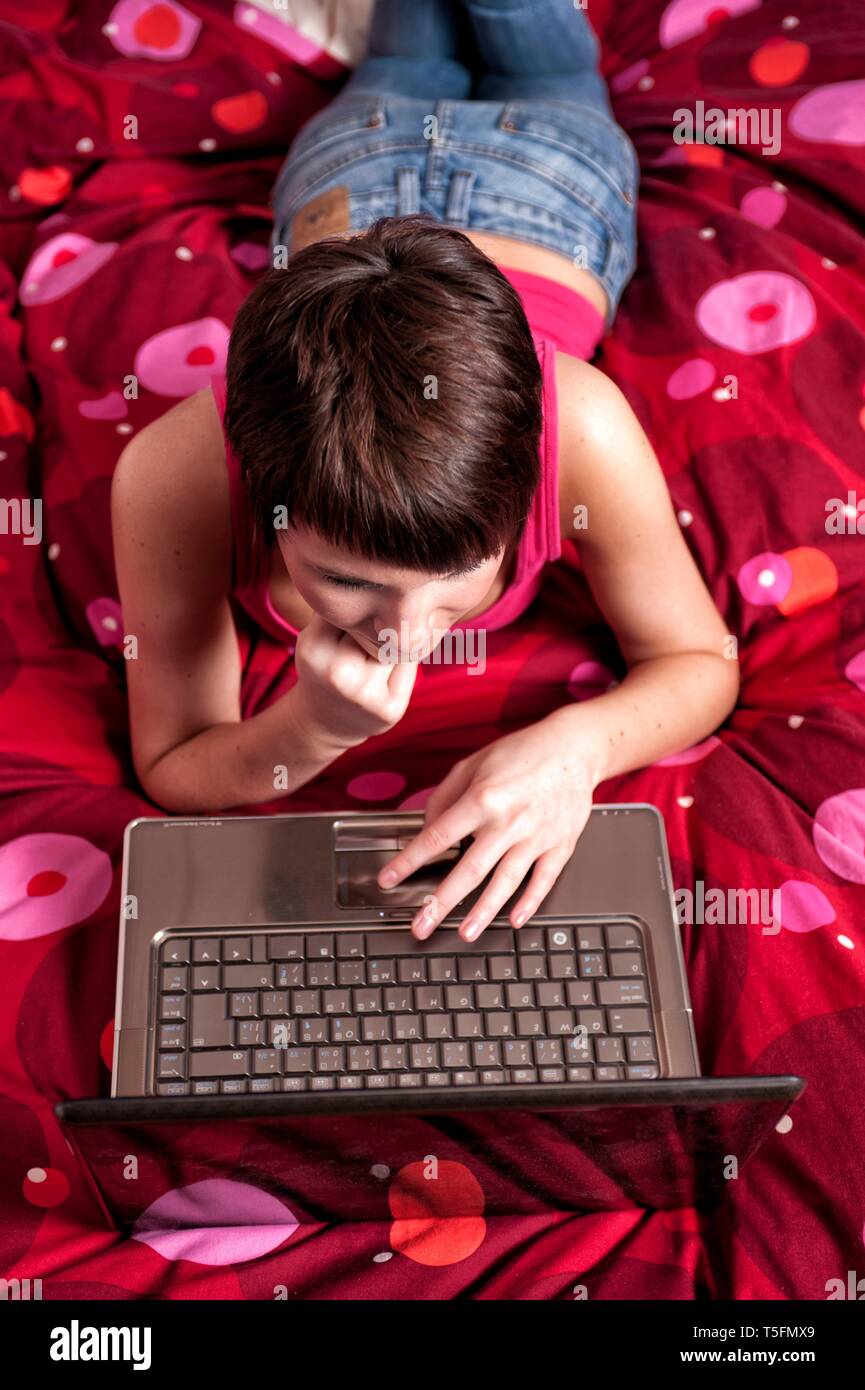 Junge Frau liegt auf ihrem Bett mit einem Leptop vor sich vor sich und schaut in die freundlich in die Kamera. Stock Photo