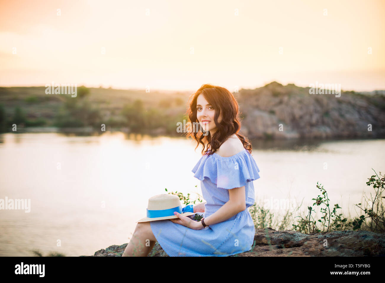 Portrait of young woman in blue romantic dress with hat sitting near river and enjoying sunset evening. Calm and harmony. Summer vacation,fun, positiv Stock Photo