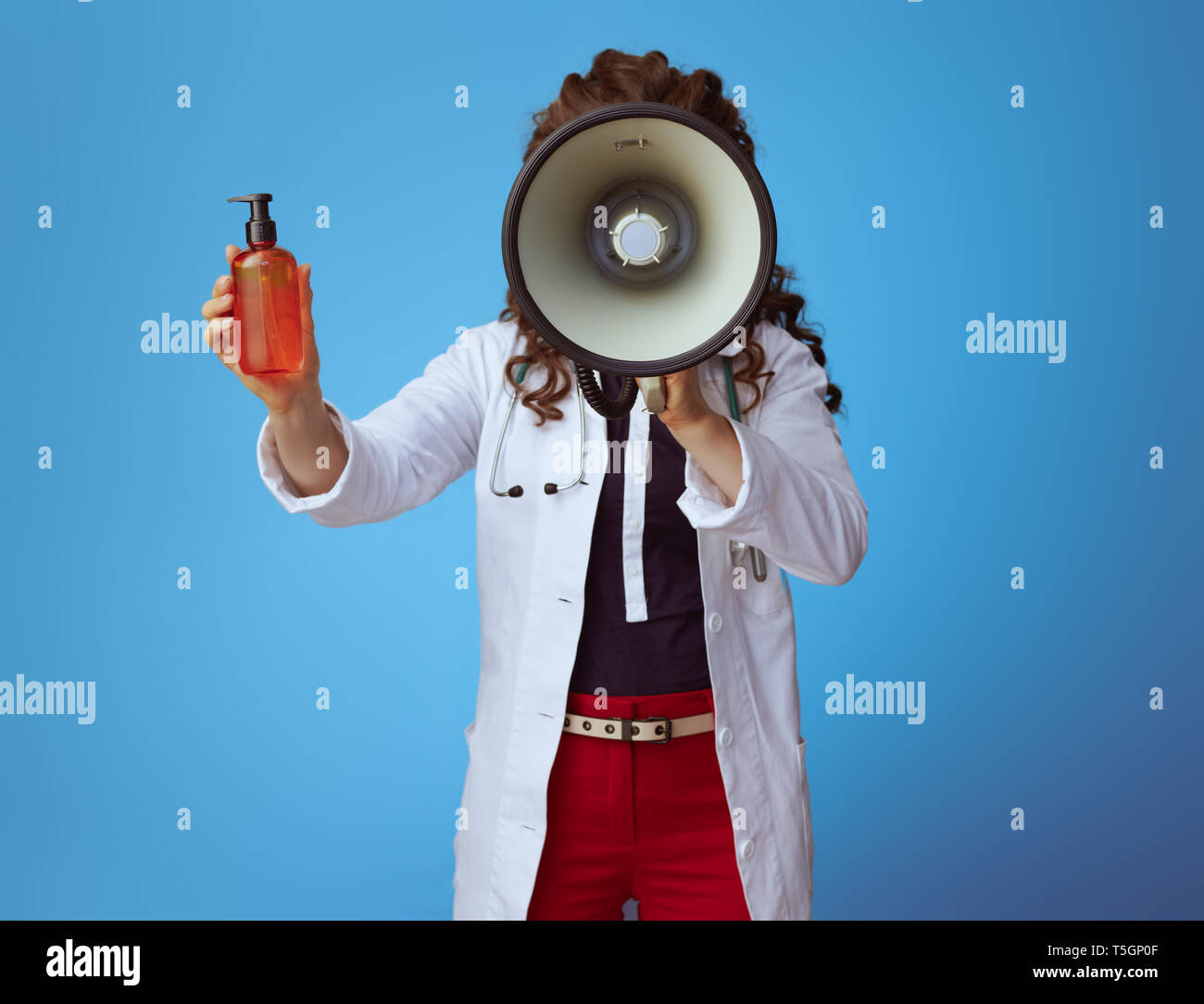 elegant medical doctor woman in bue shirt, red pants and white medical robe with megaphone showing sun cream on blue background. Stock Photo