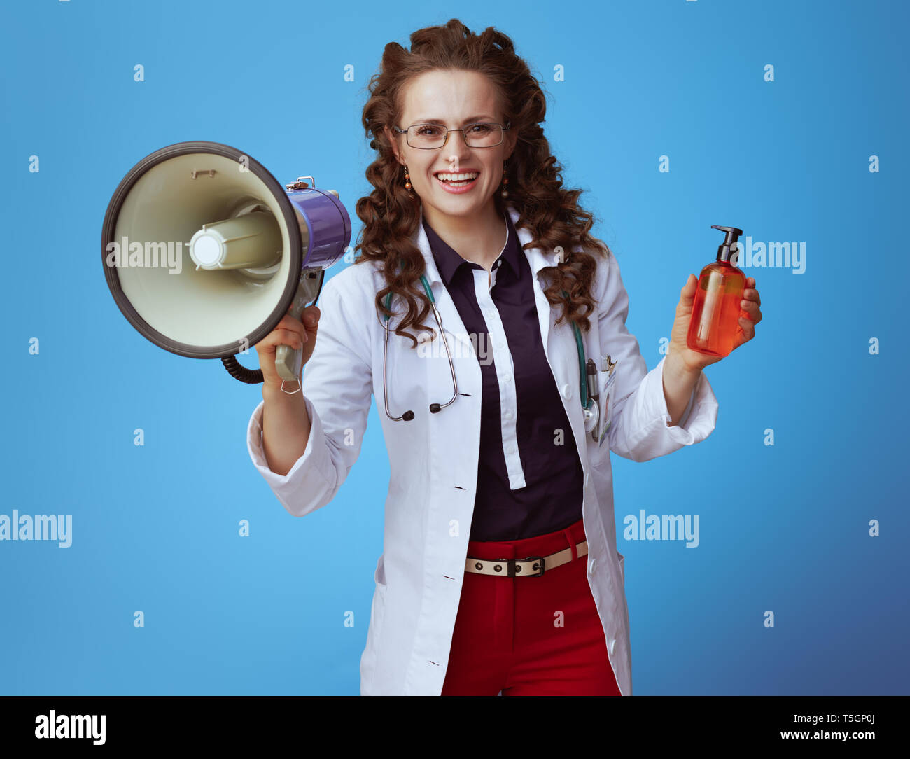 smiling modern medical practitioner woman in bue shirt, red pants and white medical robe with megaphone showing suntan lotion on blue background. Stock Photo