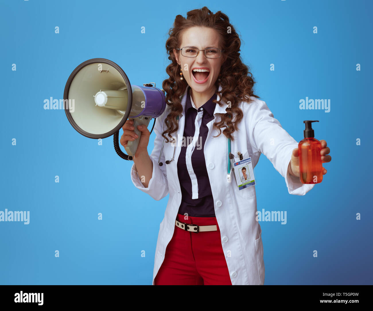 elegant medical practitioner woman in bue shirt, red pants and white medical robe with megaphone showing suntan lotion isolated on blue. Stock Photo