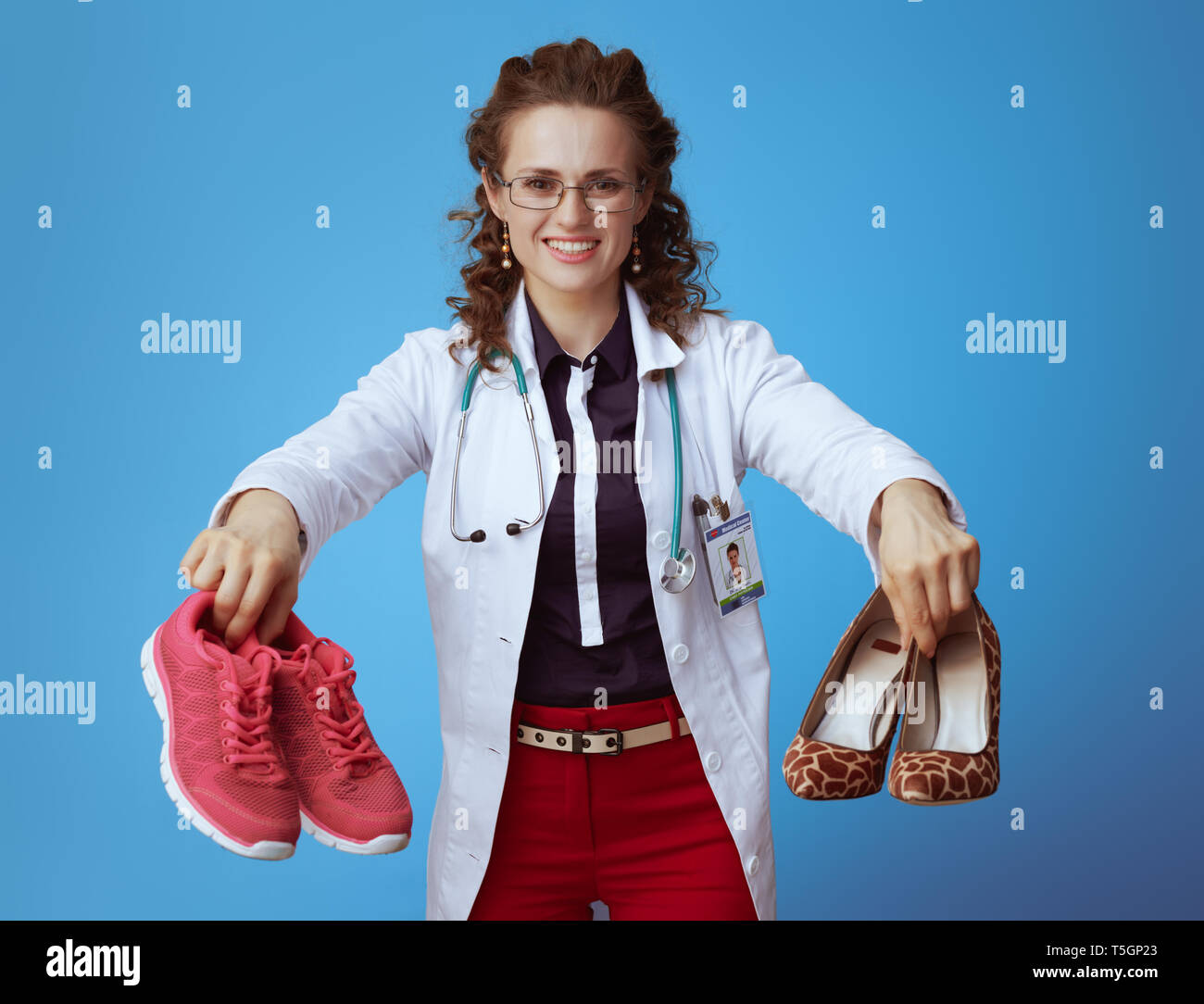 smiling modern medical practitioner woman in bue shirt, red pants and white medical robe showing fitness sneakers and high heel shoes against blue bac Stock Photo