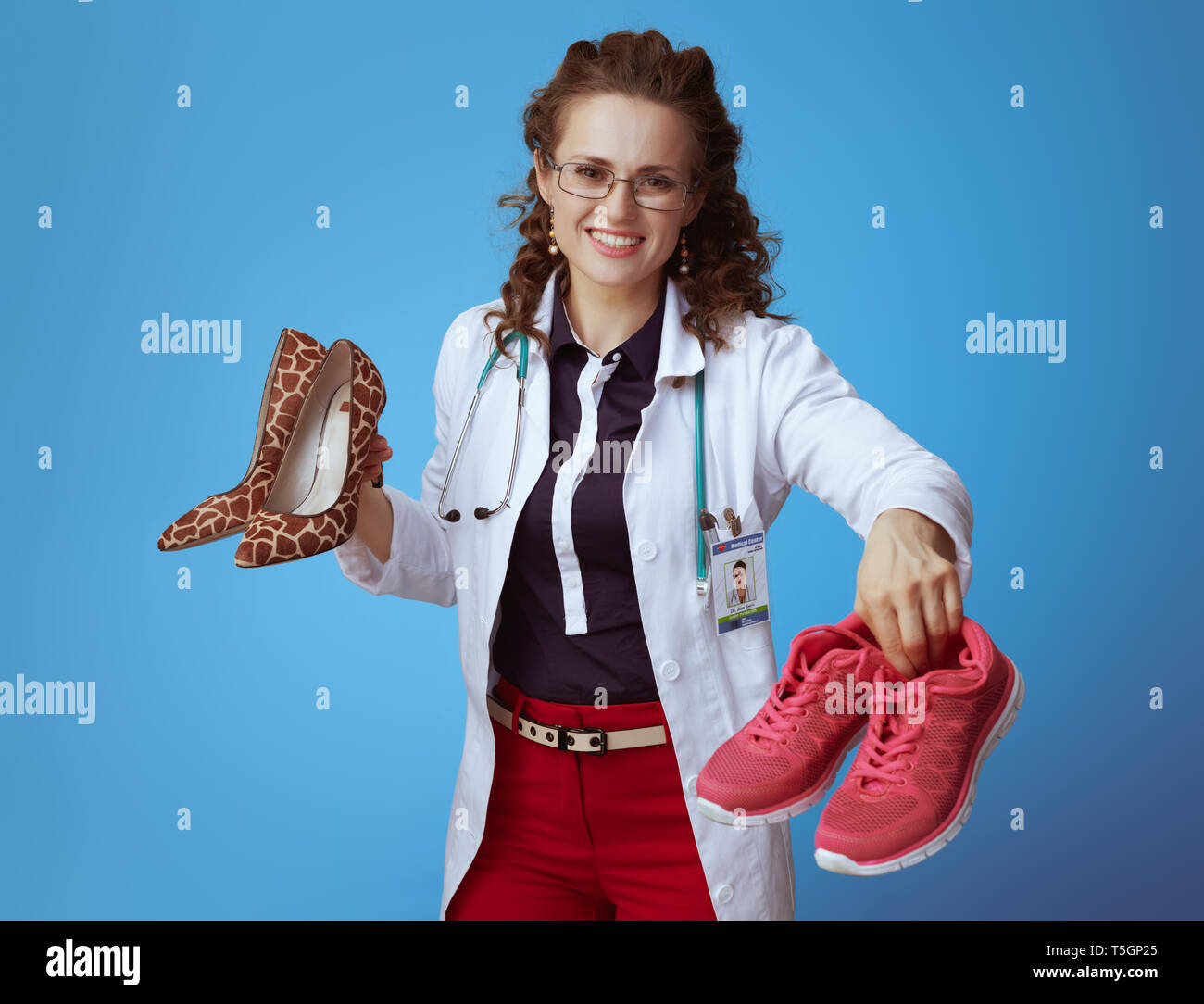 happy elegant physician woman in bue shirt, red pants and white medical robe giving fitness sneakers while holding high heel shoes in other hand isola Stock Photo