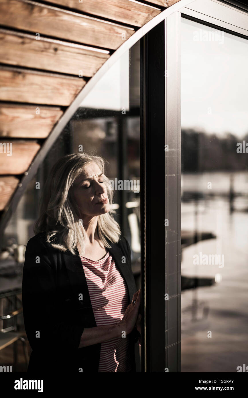 Businesswoman standing on a houseboat,enjoying the sun at the window with eyes closed Stock Photo
