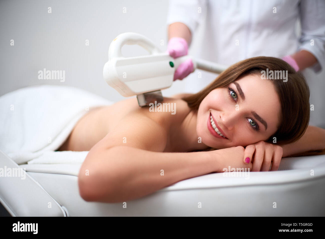Young pretty smiling woman back laser epilation in beauty salon. Cosmetologist doing depilation treatment with elos device. Beautician hands in gloves Stock Photo