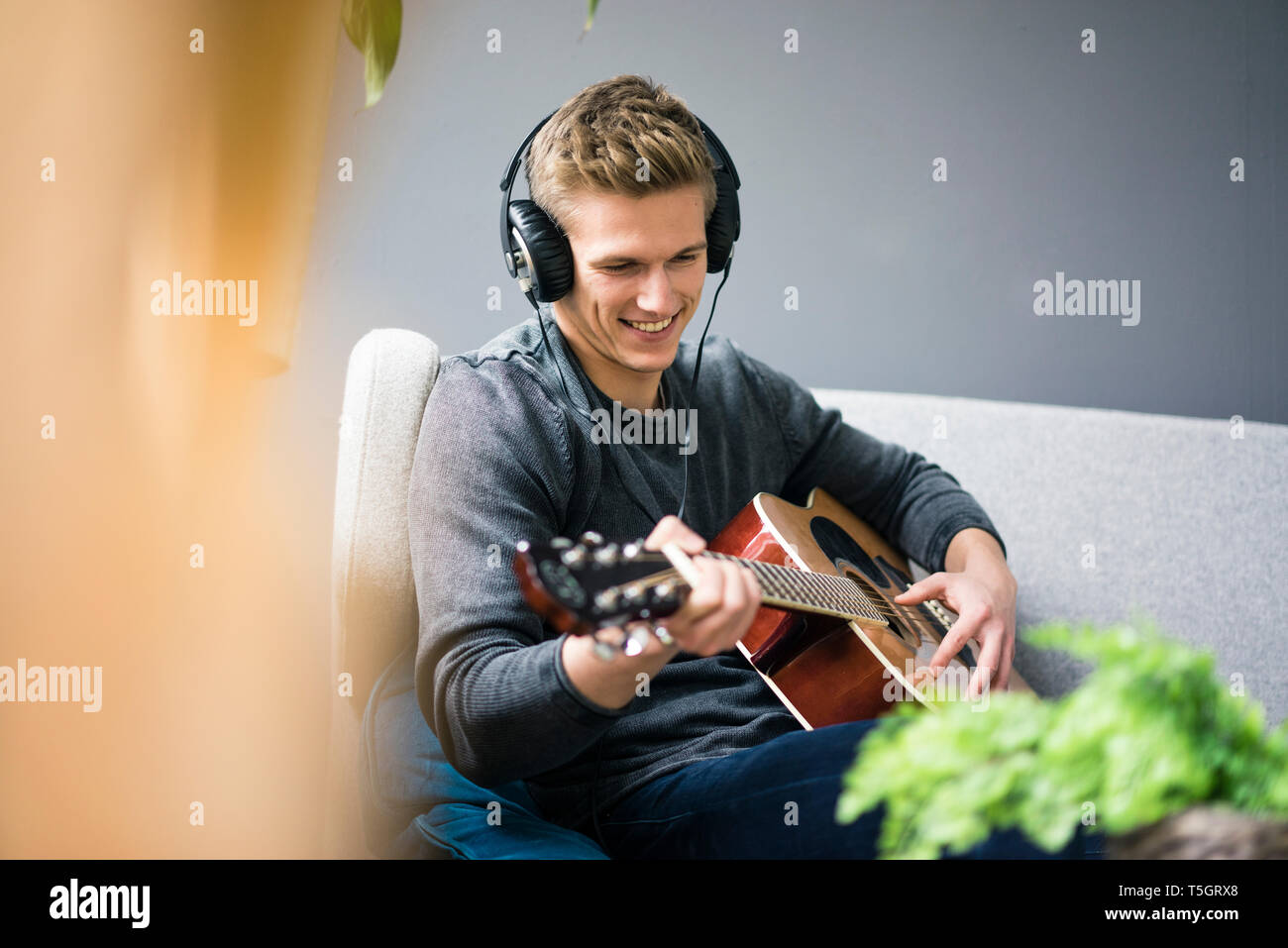 Smiling young man with headphones sitting on couch playing guitar Stock Photo