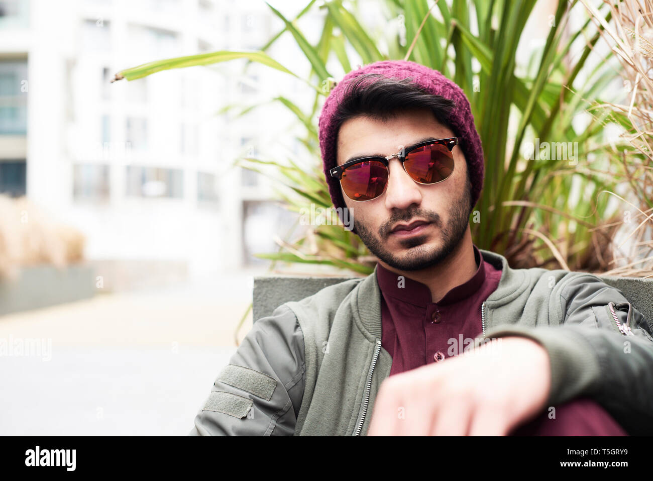 Portrait of fashionable man in mirrored colored sunglasses and purple knit hat against green plant Stock Photo