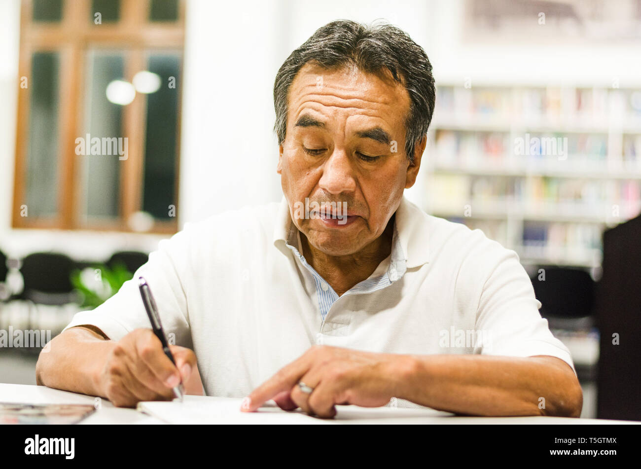 Serious senior man sitting on a library bench writing in his book. Senior man sitting in a university classroom. Stock Photo