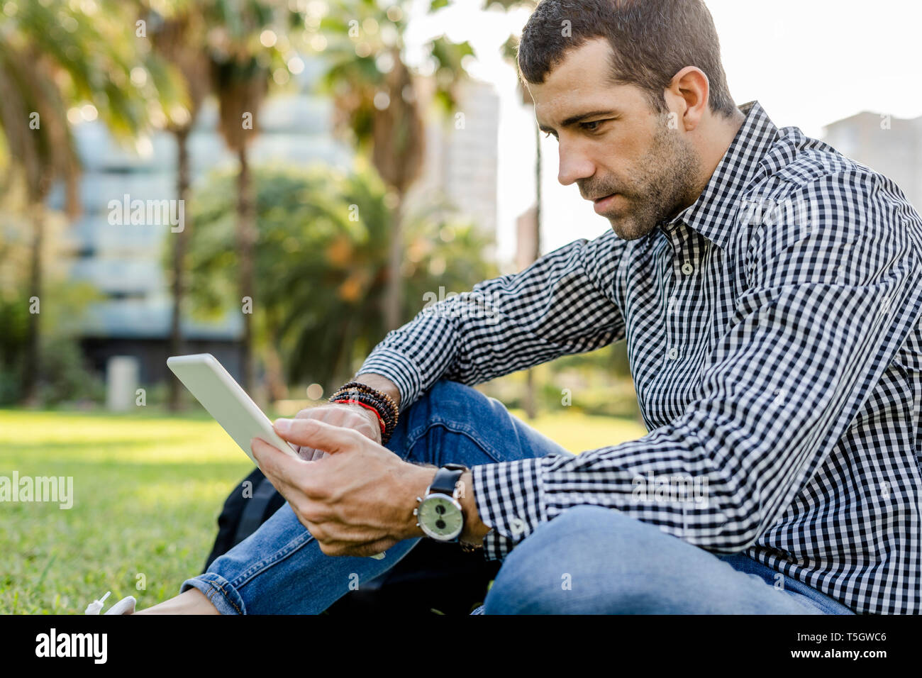 Man sitting on meadow in city park looking at digital tablet Stock Photo
