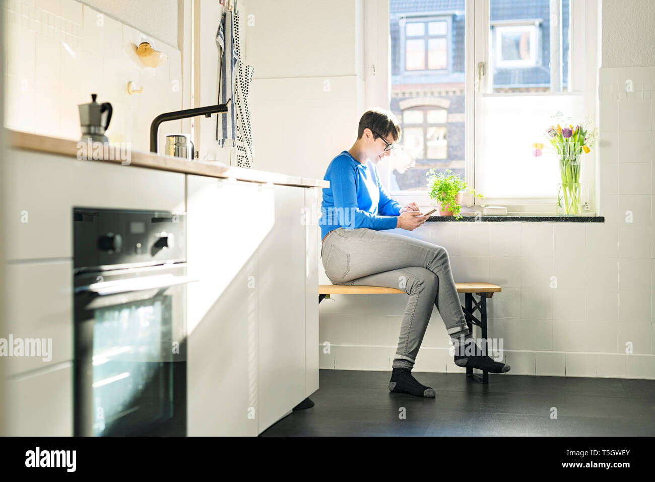 Short-haired woman sitting on bench in kitchen at the window using smartphone Stock Photo