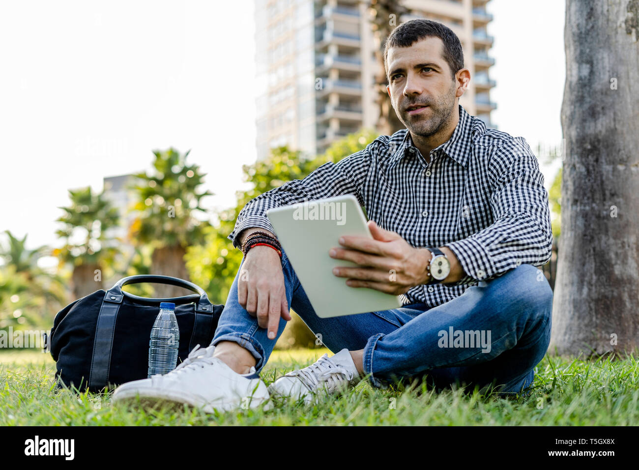 Portrait of man with digital tablet sitting on meadow in city park thinking Stock Photo