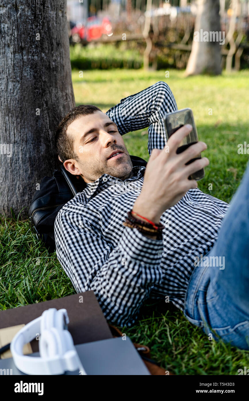Portrait of man lying on meadow in city park looking at cell phone Stock Photo
