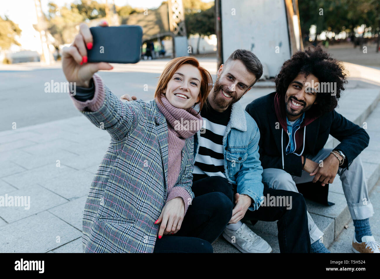 Three happy friends sitting outdoors taking a selfie Stock Photo