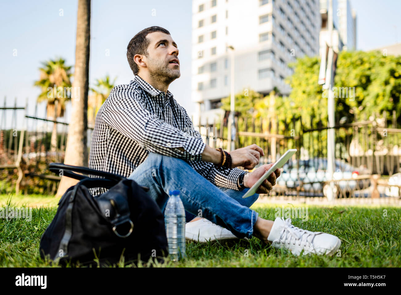 Man sitting on meadow in city park with digital tablet looking up Stock Photo
