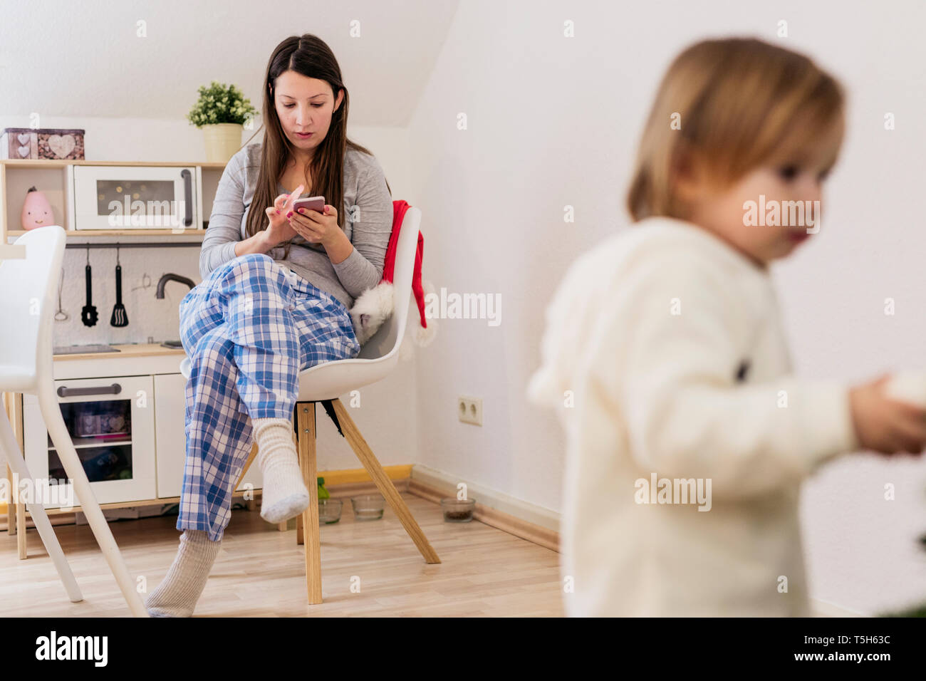 Woman sitting in kitchen using smartphone while little daughter standing in the foreground Stock Photo