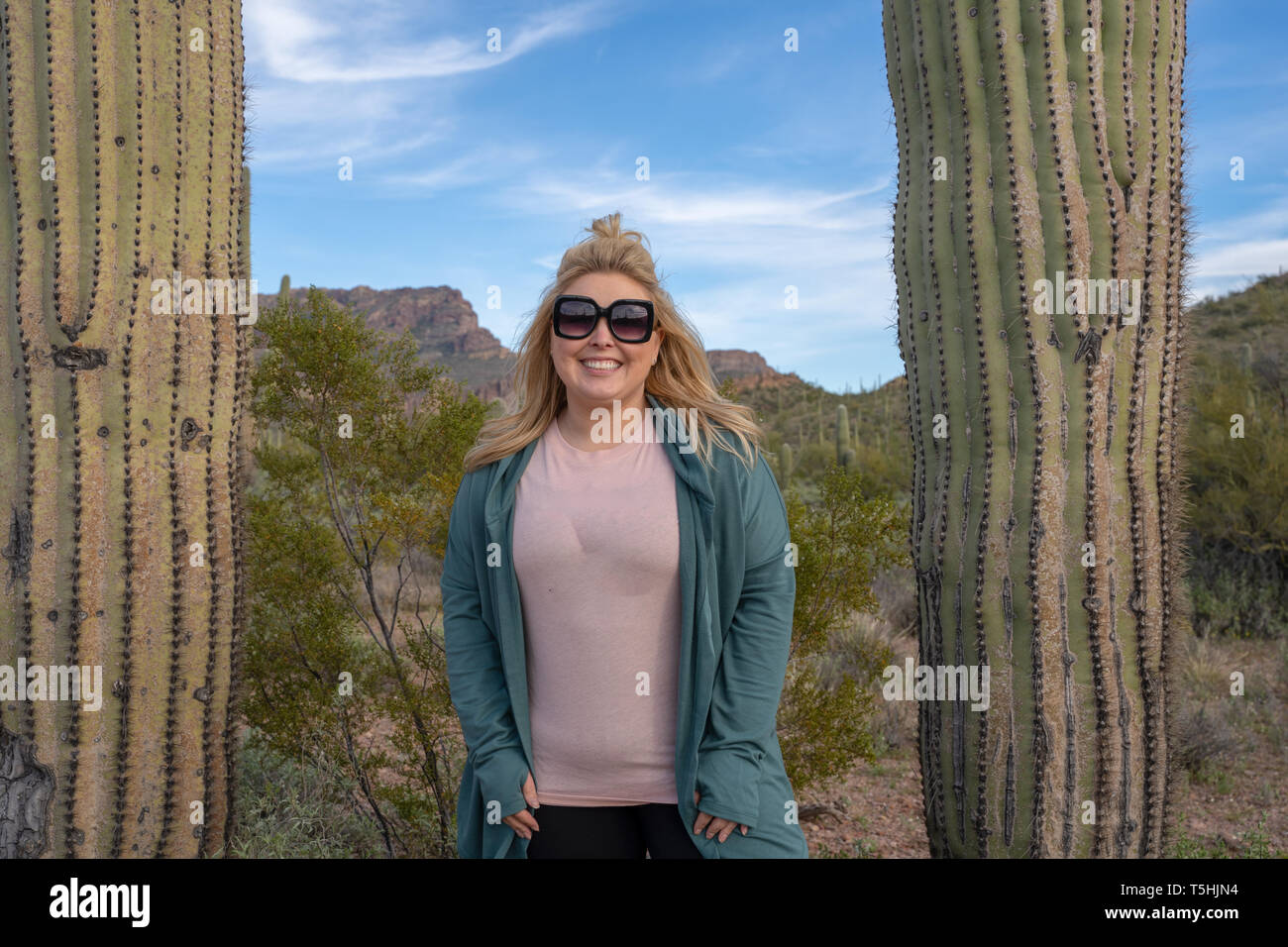 Happy, relaxed blond woman wearing sunglasses stands between two saguaro cactus (cacti) in Arizona, wearing casual clothing Stock Photo