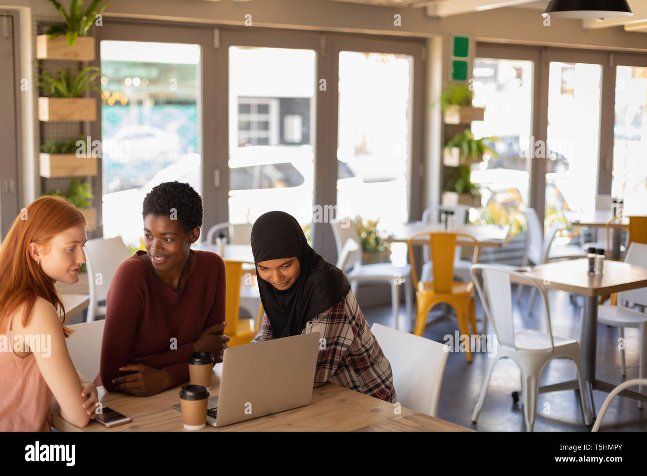 Female friends interacting with each other while using laptop in a cafe Stock Photo