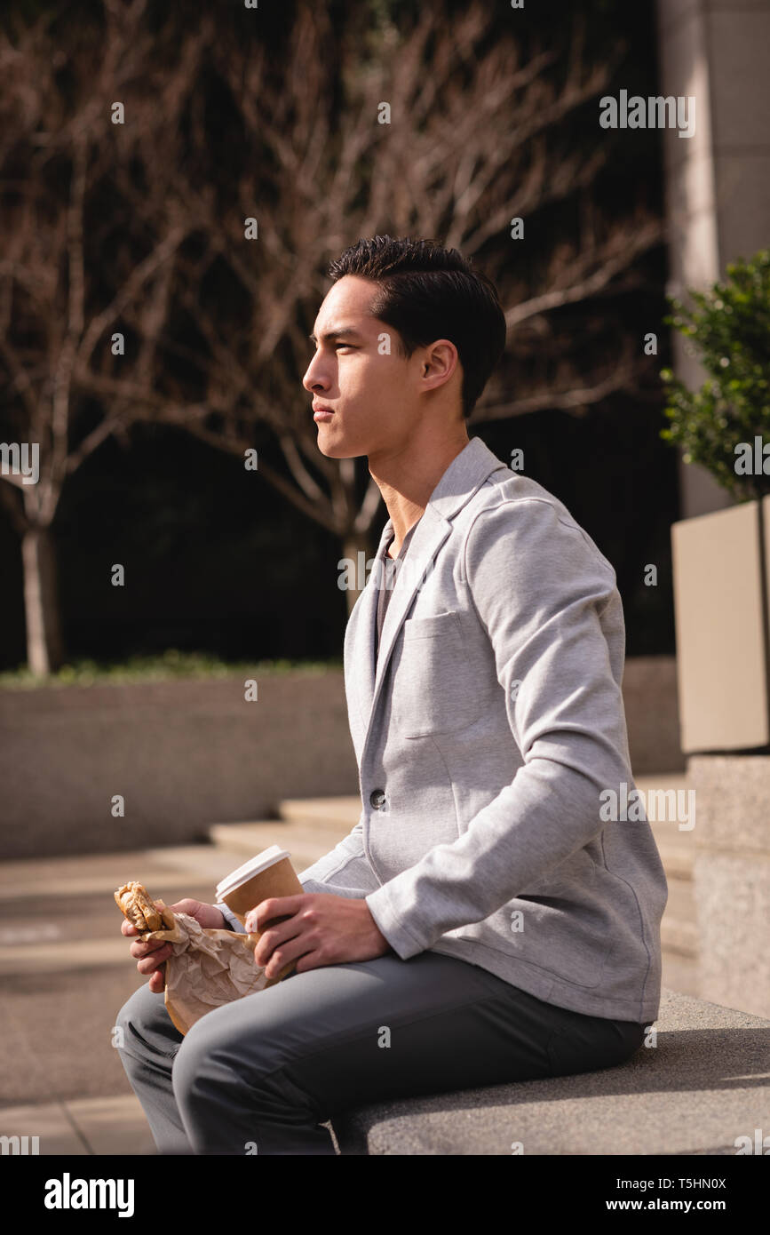 Young man having coffee and breakfast while sitting on stone bench Stock Photo