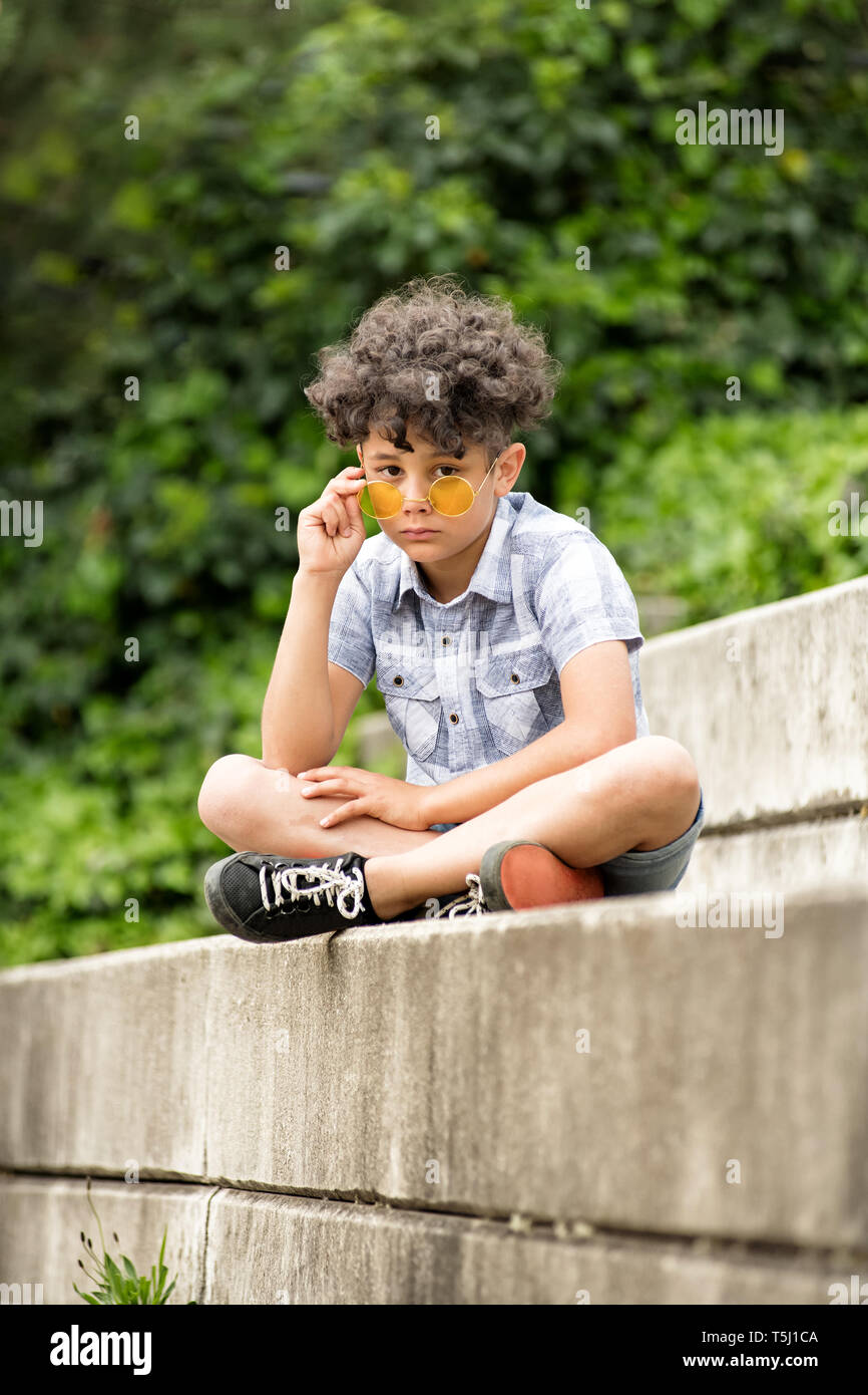Solemn little boy wearing yellow sunglasses seated on a concrete step outdoors lowering the glasses to peer over at the camera Stock Photo