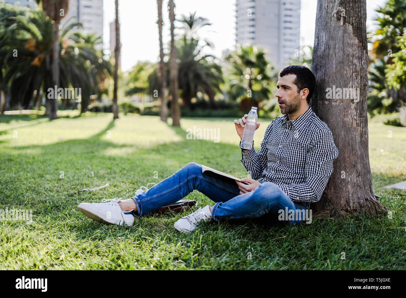 Man on a meadow in city park leaning against tree drinking water Stock Photo