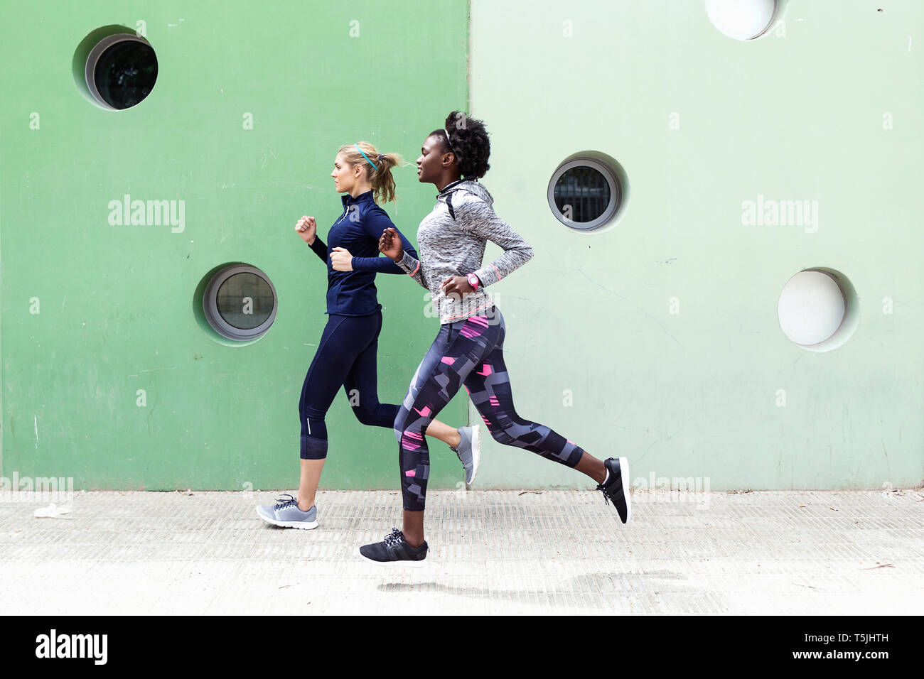 Two sporty young women running together in the city passing wall with round windows Stock Photo