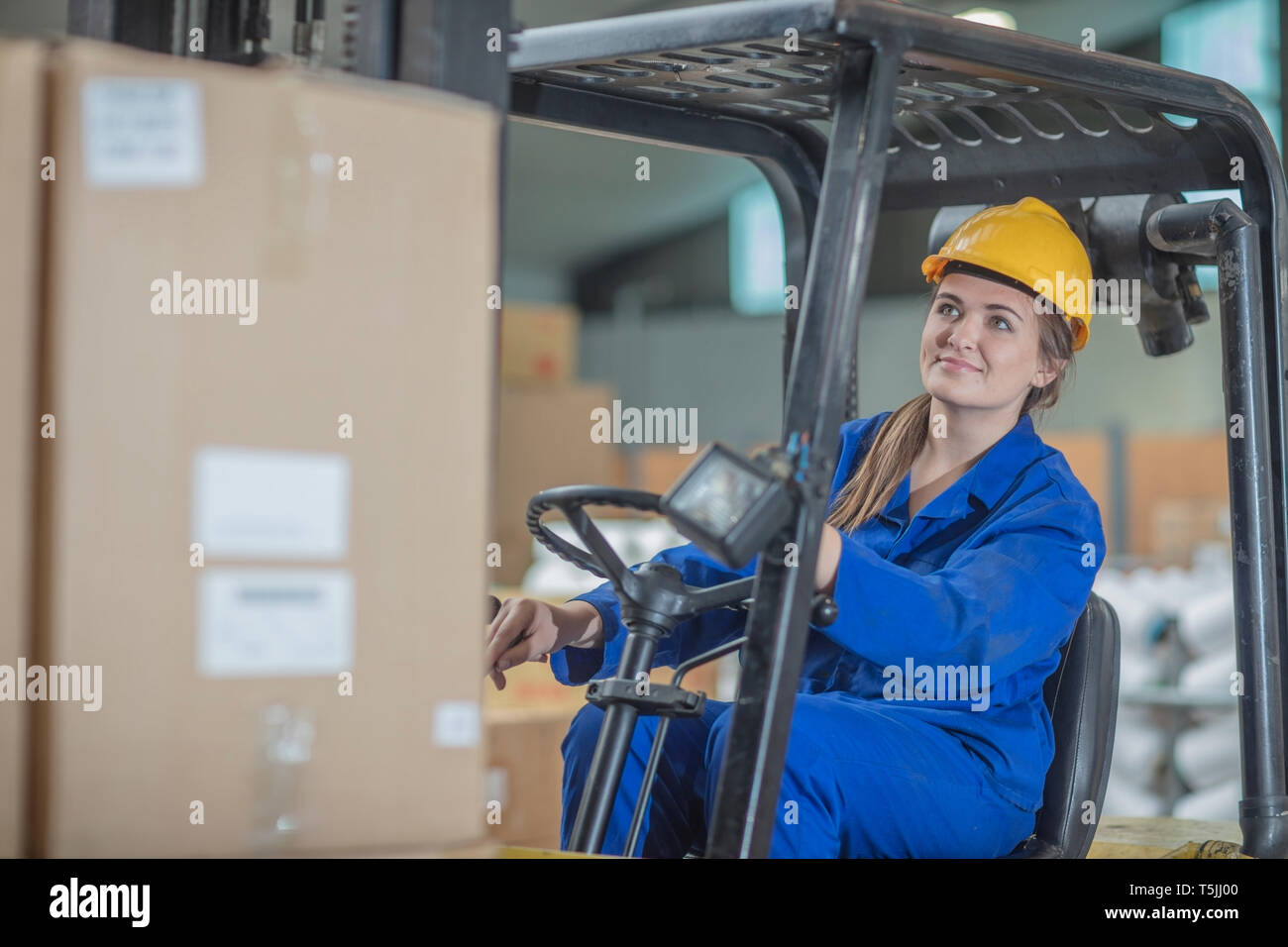 Smiling woman wearing hard hat driving forklift in factory Stock Photo