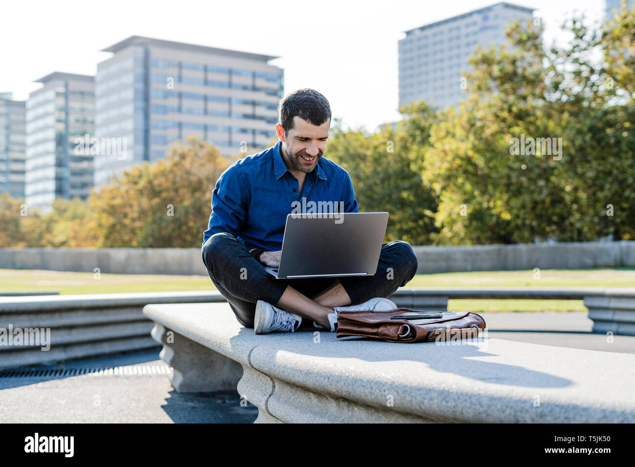 Smiling businessman sitting on bench outdoors working on laptop Stock Photo