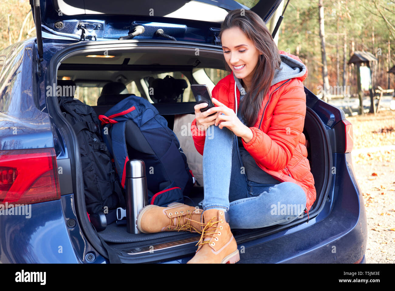 Woman using smartphone while sitting in car trunk during road trip Stock Photo