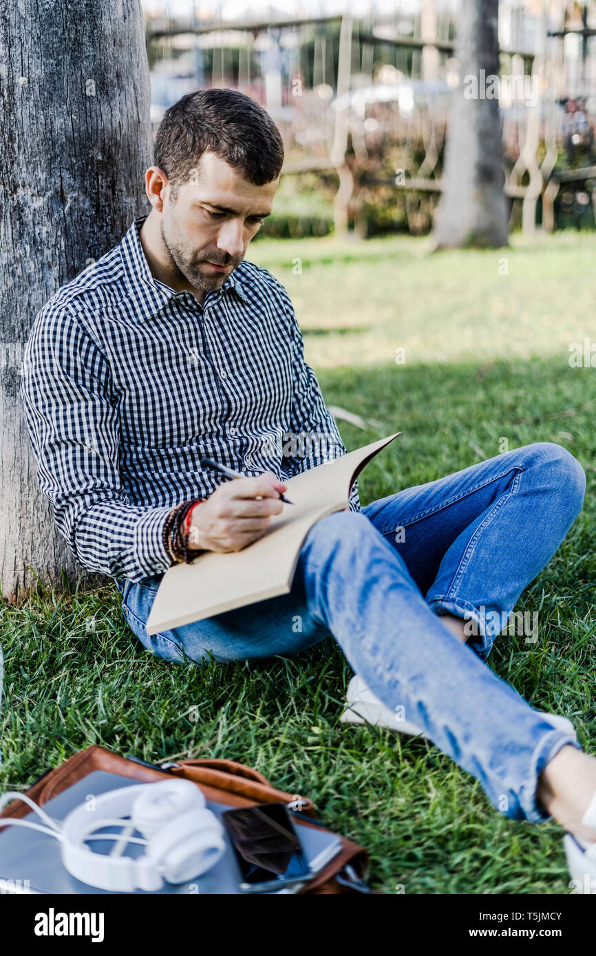Man on a meadow leaning against tree trunk taking notes Stock Photo