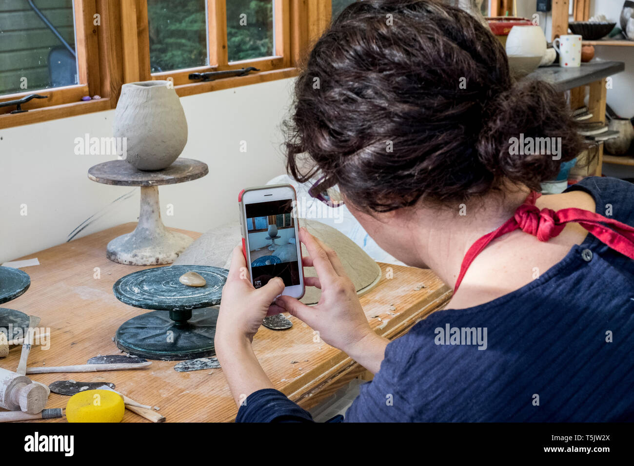 Over the shoulder view of woman sitting in her ceramics workshop, checking her mobile phone. Stock Photo