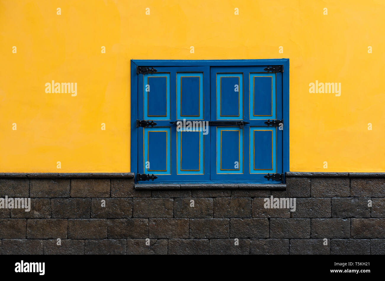 A colourful closed blue window with yellow wall and black stone bricks underneath in the historic city center of Cuenca, Ecuador. Stock Photo