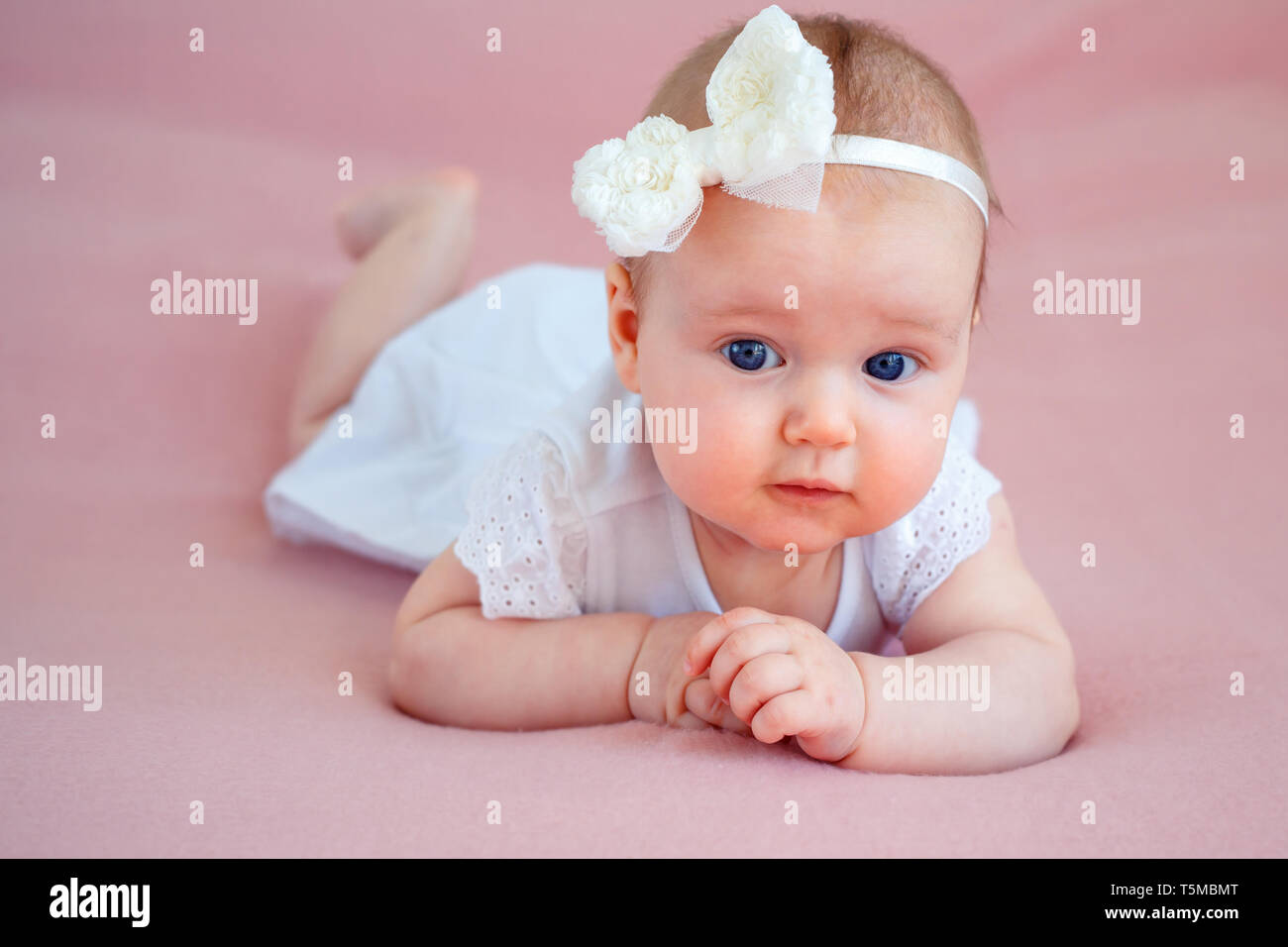 Portrait of a cute newborn baby girl in white dress and hairband lying on her stomach in bedroom on pink soft blanket Stock Photo